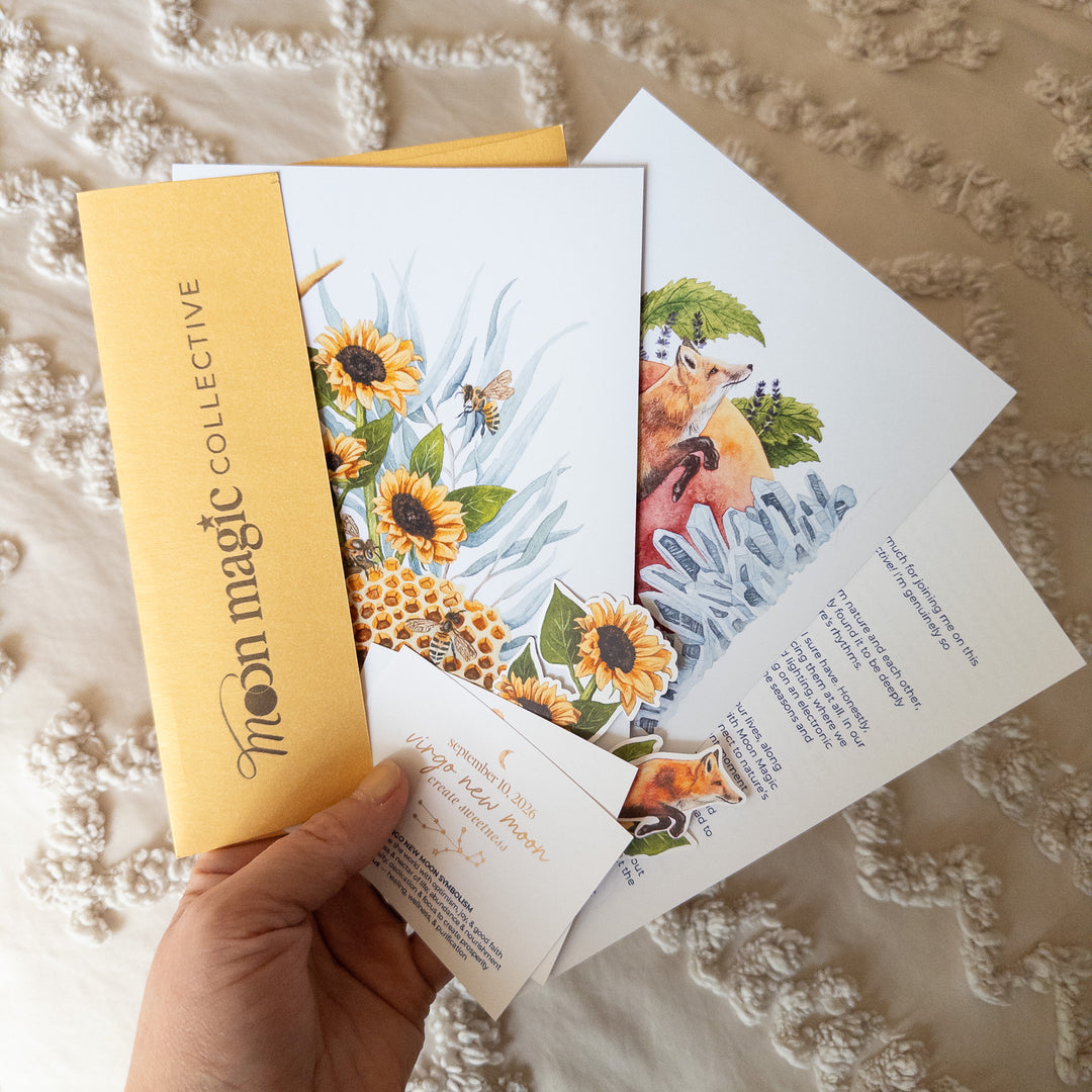 A hand holds a set of printed 5x7 fine art prints and a gold envelope from Moon Magic Collective, featuring whimsical cosmic watercolor illustrations of moons, sunflowers, a fox, and bees, laid over a textured cream fabric background.